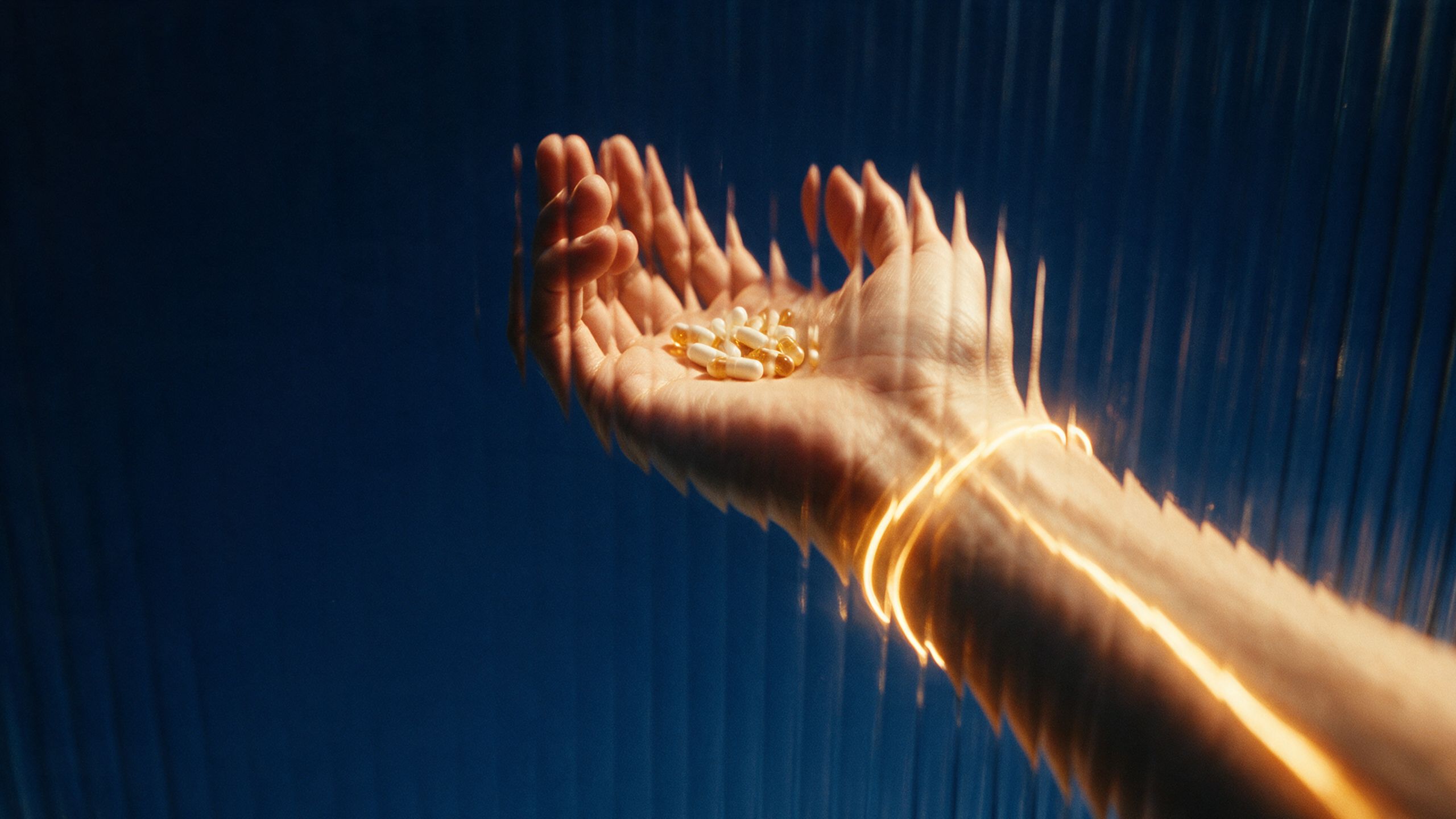 An outstretched palm holding a few capsules, seen through ribbed glass against a deep blue background, with a ribbon of golden light wrapping the wrist